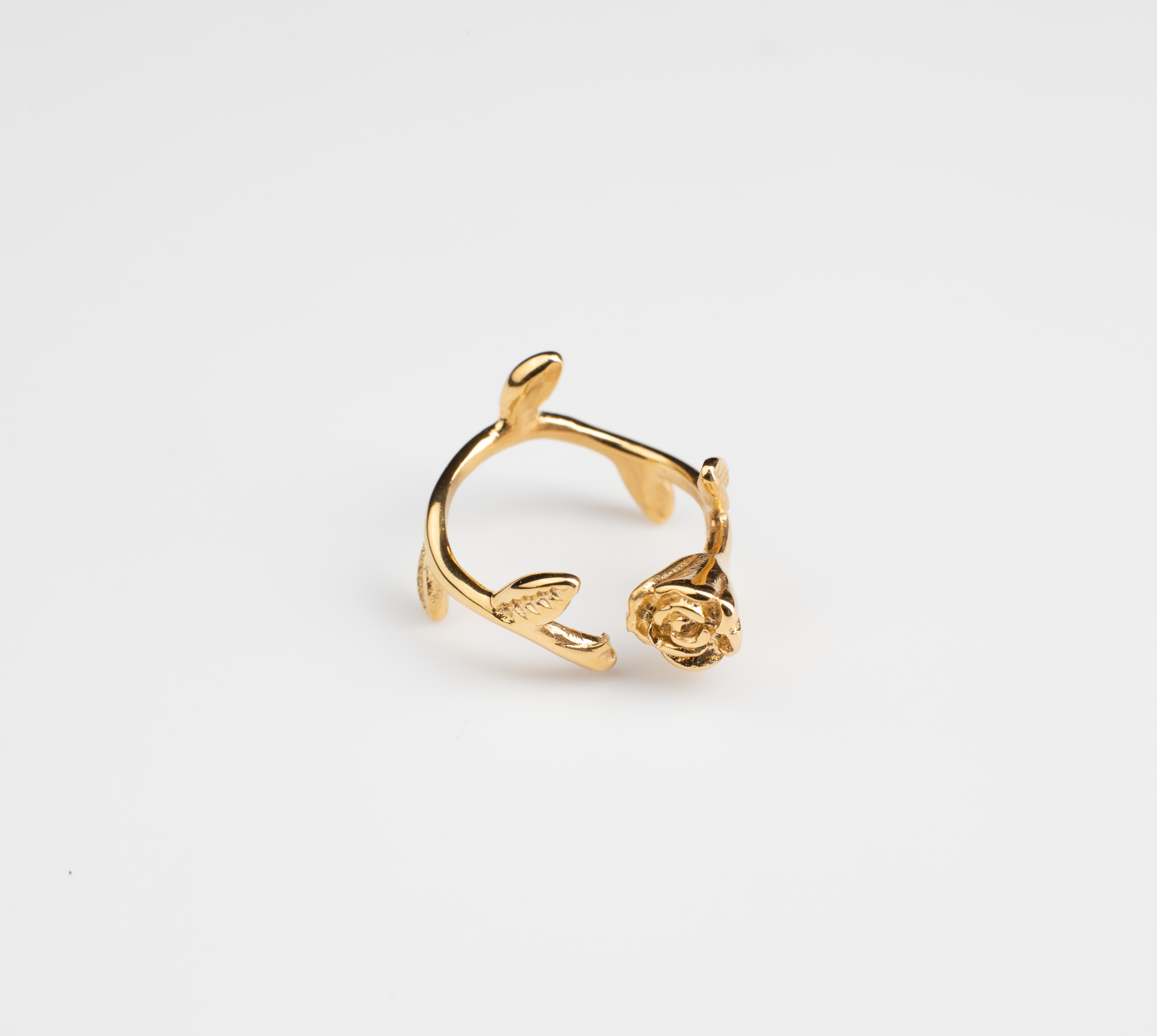 This ring features a shiny gold tone throughout, with an open band design allowing for adjustable sizing. The band is adorned with extended leaves, and at one end of the band sits a intricately sculpted, lifelike rose flower.