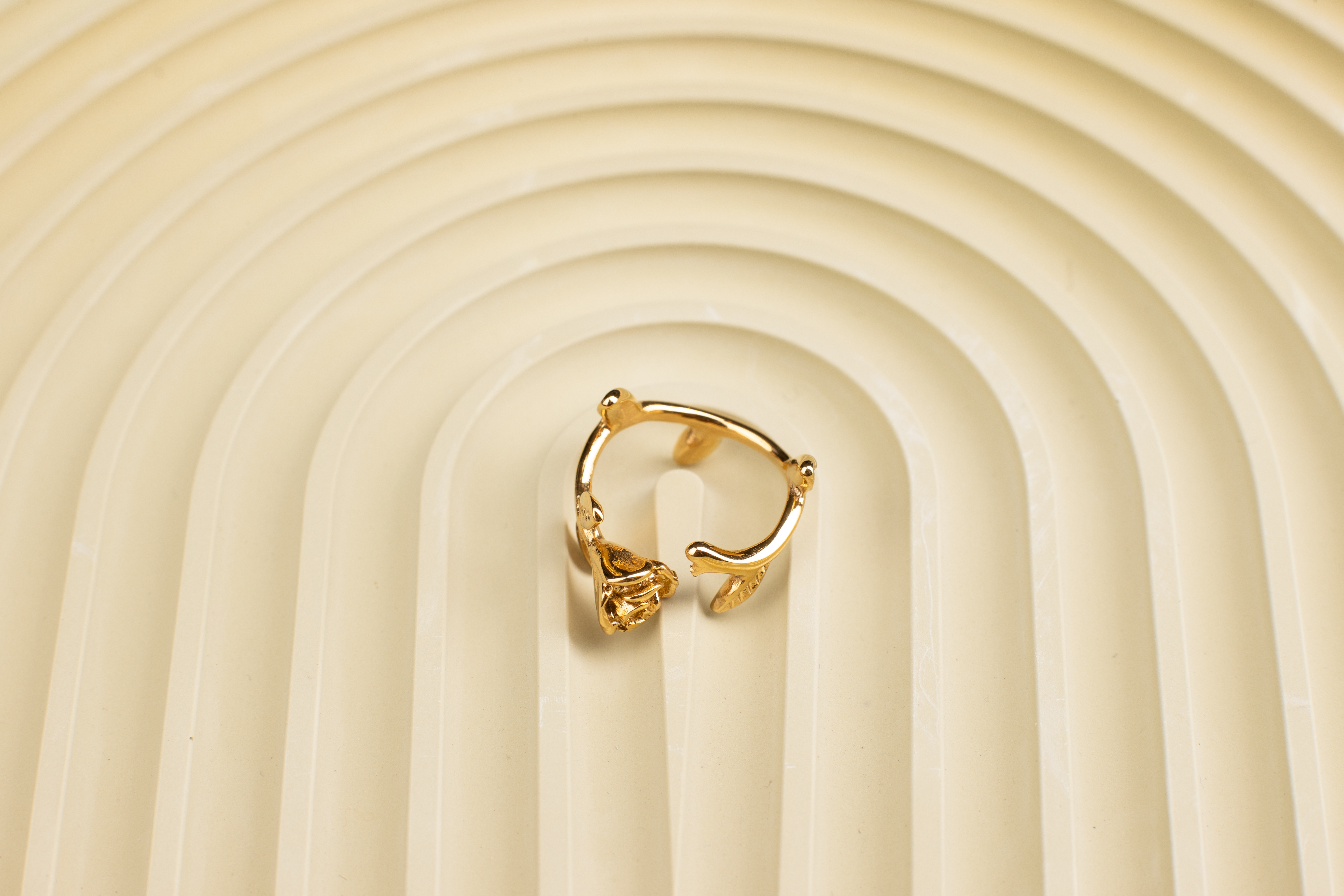 This ring features a shiny gold tone throughout, with an open band design allowing for adjustable sizing. The band is adorned with extended leaves, and at one end of the band sits a intricately sculpted, lifelike rose flower.