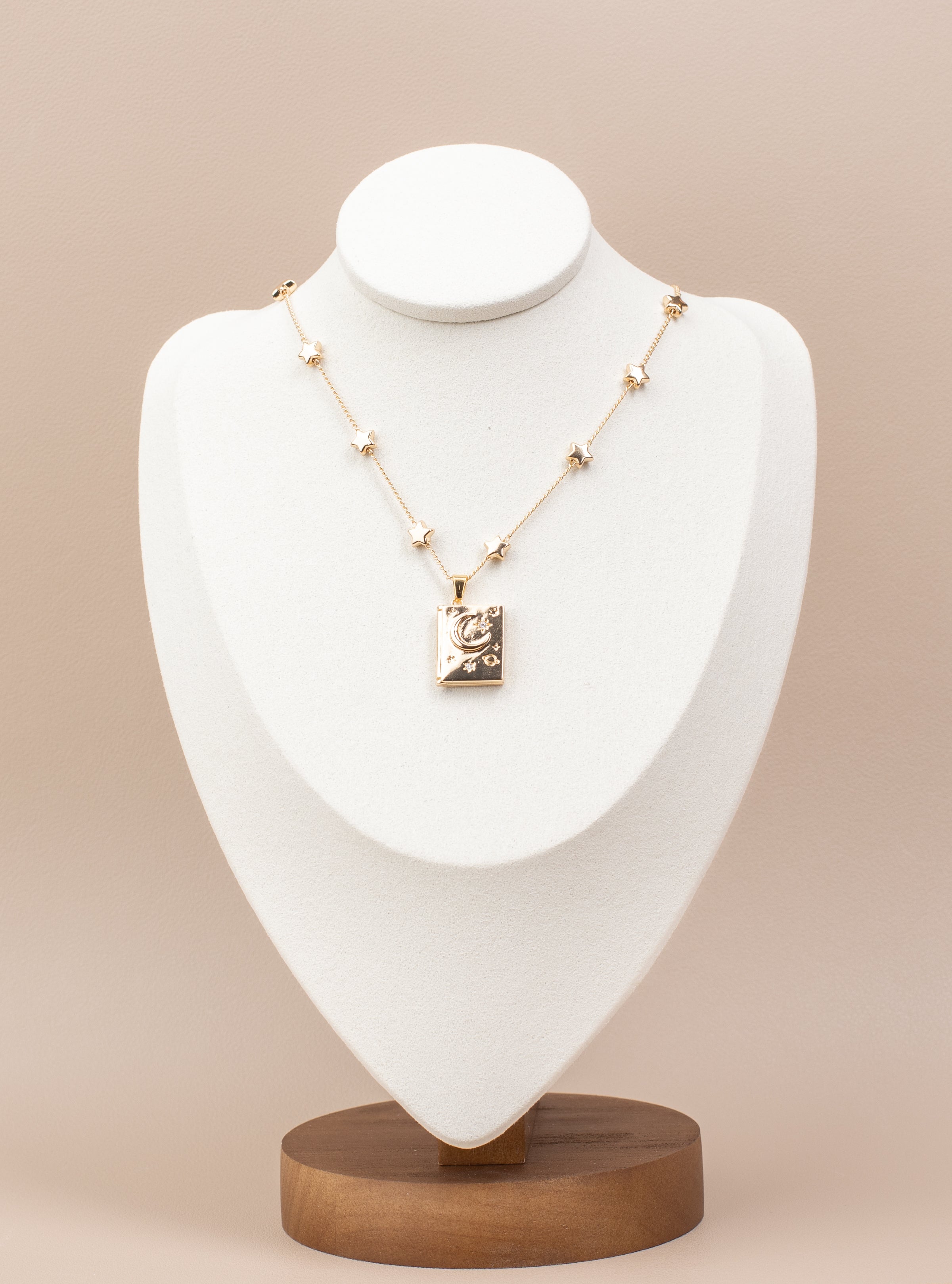 Book-shaped locket necklace with a celestial design featuring stars, a crescent moon, and sparkling gemstone accents. Made from copper with 14K gold plating, this openable locket is elegantly displayed on a white velvet bust stand with a wooden base, perfect for showcasing its timeless charm and photo-holding functionality.