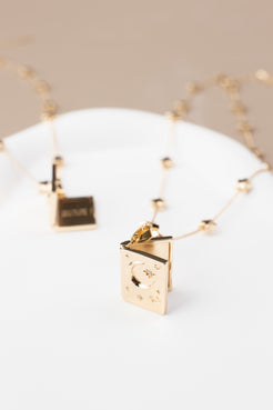 Book-shaped locket necklace with a celestial design, featuring stars, a crescent moon, and sparkling gemstone accents. Made from copper with 14K gold plating, the locket is shown partially open on a soft beige background, highlighting its elegant design and photo-holding functionality.