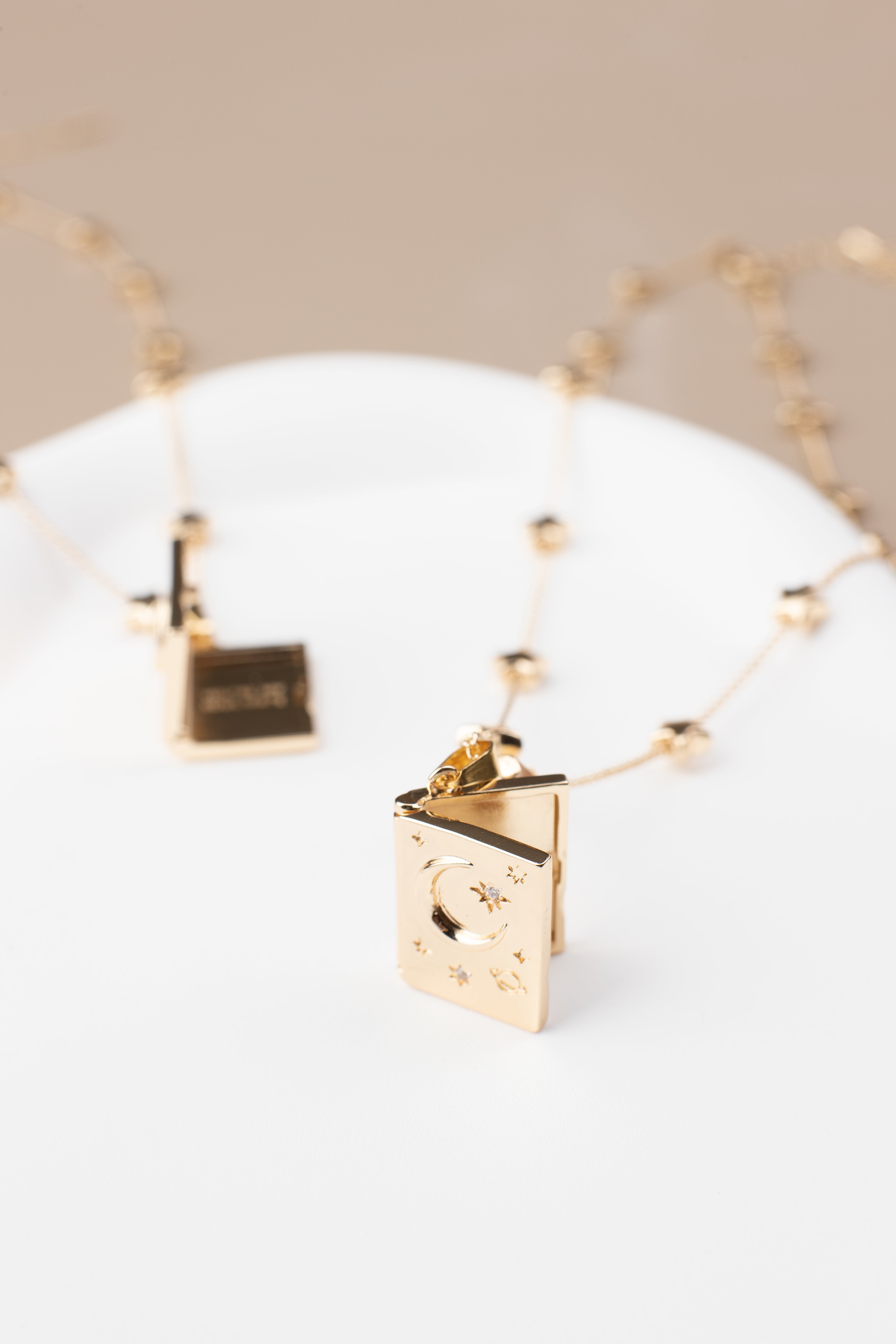 Book-shaped locket necklace with a celestial design, featuring stars, a crescent moon, and sparkling gemstone accents. Made from copper with 14K gold plating, the locket is shown partially open on a soft beige background, highlighting its elegant design and photo-holding functionality.