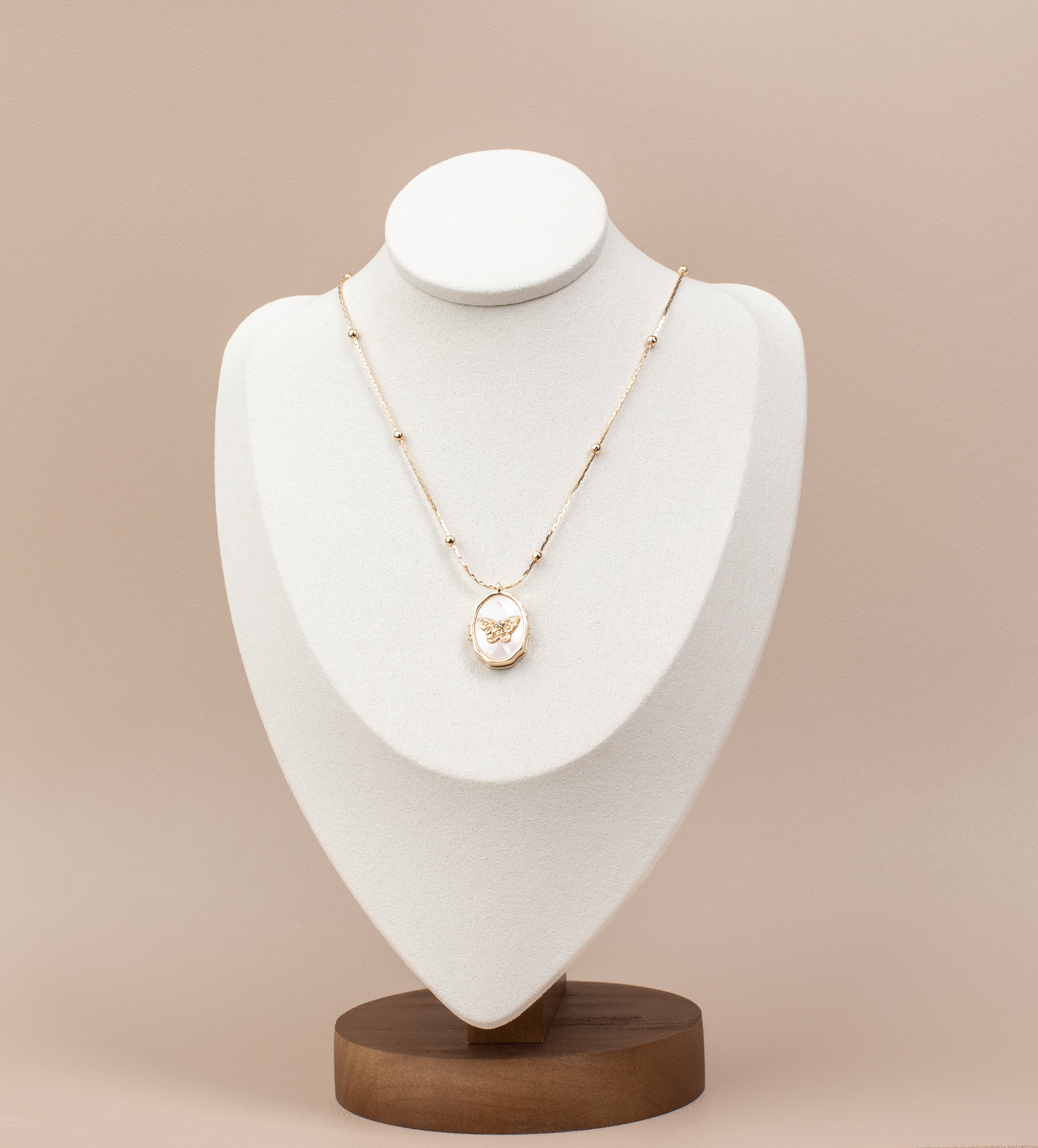 Oval locket necklace featuring a butterfly design over a mother-of-pearl background, crafted from copper with 14K gold plating. Displayed elegantly on a white velvet bust stand with a wooden base, highlighting its delicate and timeless design.
