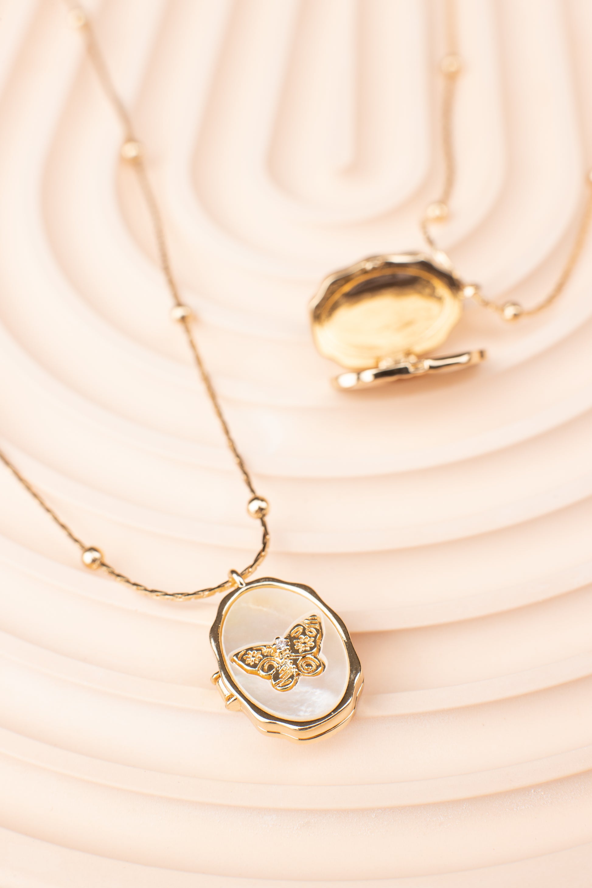 Oval locket necklace with a butterfly design over a mother-of-pearl background, crafted from copper with 14K gold plating. The locket is shown partially open, revealing its hidden photo compartment. Displayed on a soft beige textured surface, emphasizing its elegant and functional design.