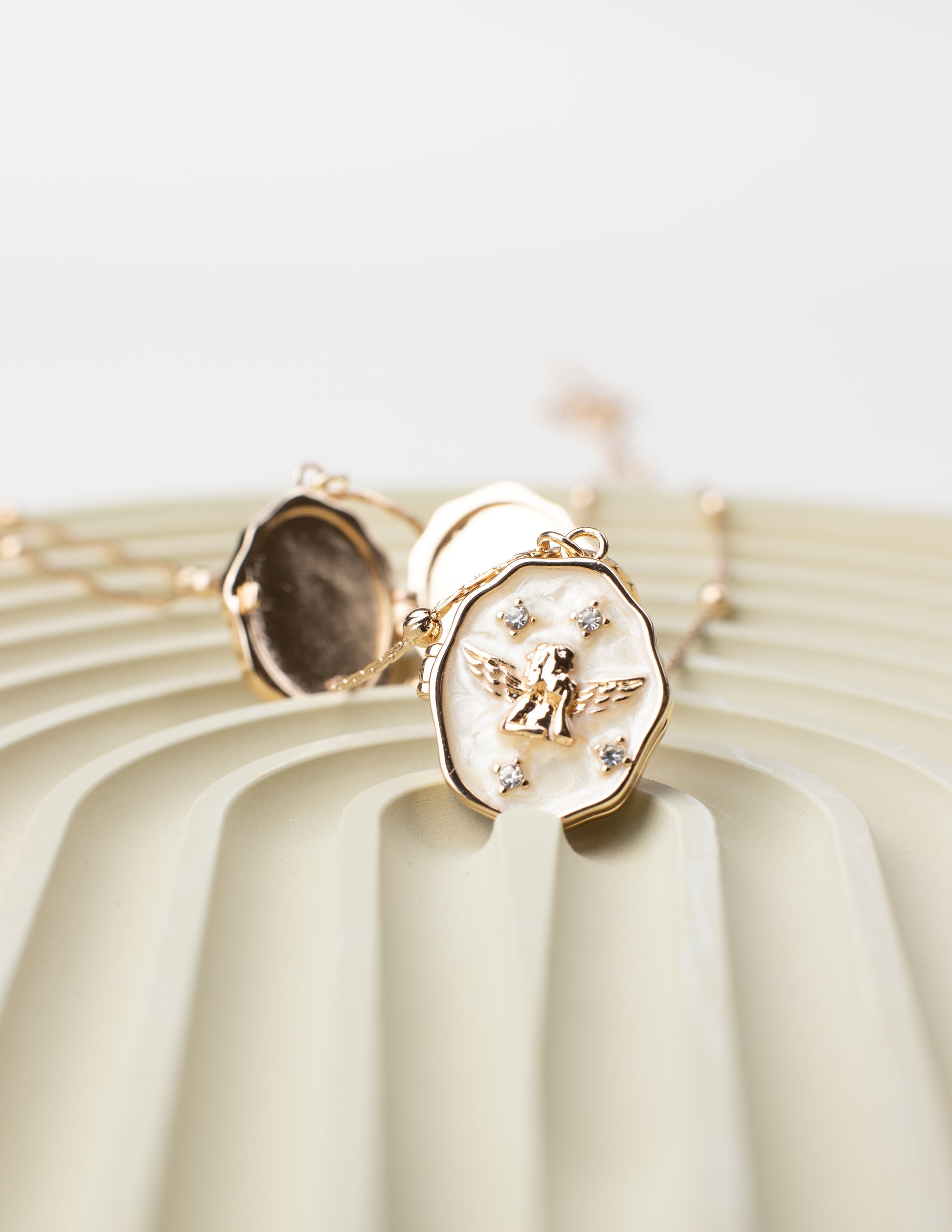 An oval locket necklace with an angel design and shimmering gemstone embellishments, crafted from 18K gold-plated copper. The locket is partially open, showcasing its photo-holding compartment, beautifully displayed on a textured, pastel-toned surface to emphasize its intricate details and elegant craftsmanship.