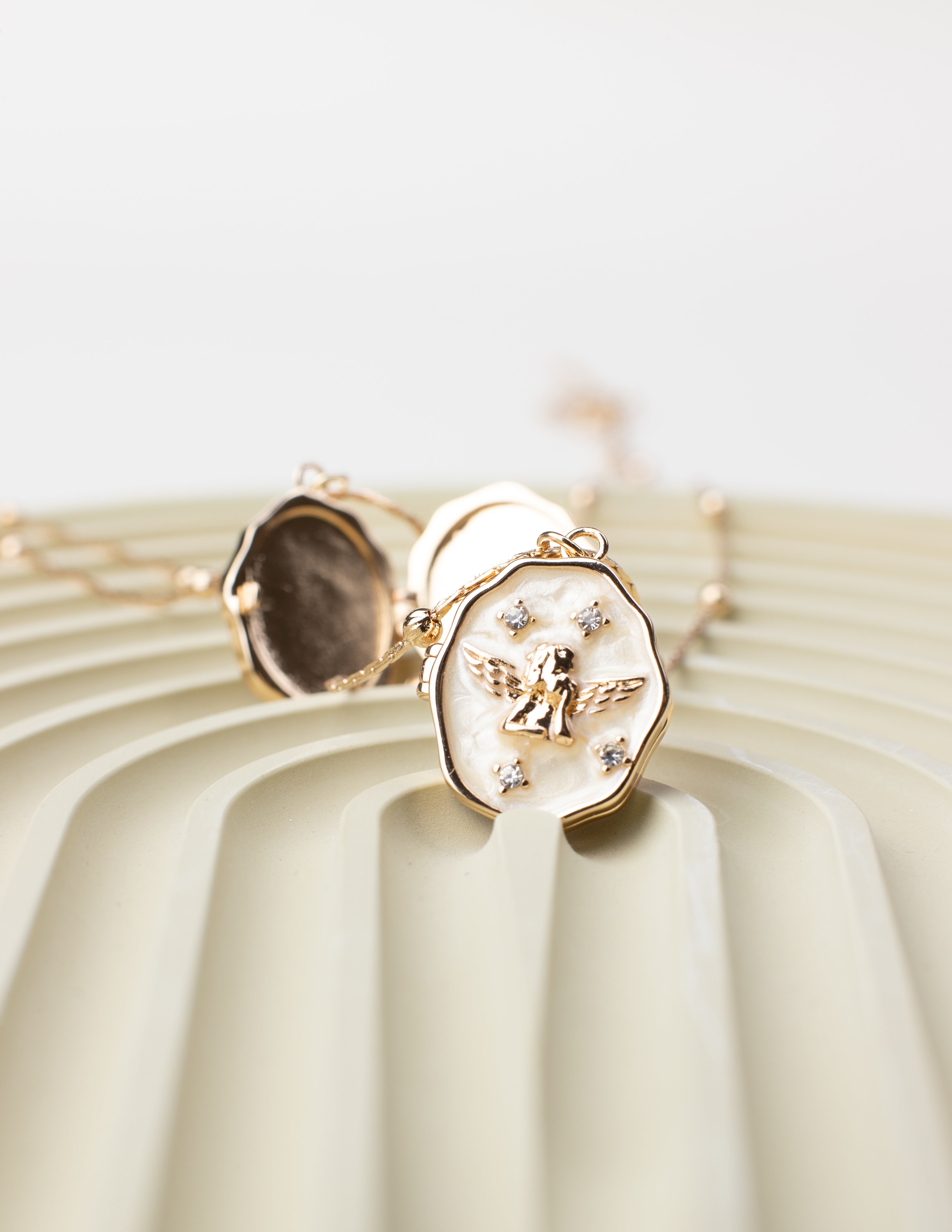 An oval locket necklace with an angel design and shimmering gemstone embellishments, crafted from 18K gold-plated copper. The locket is partially open, showcasing its photo-holding compartment, beautifully displayed on a textured, pastel-toned surface to emphasize its intricate details and elegant craftsmanship.