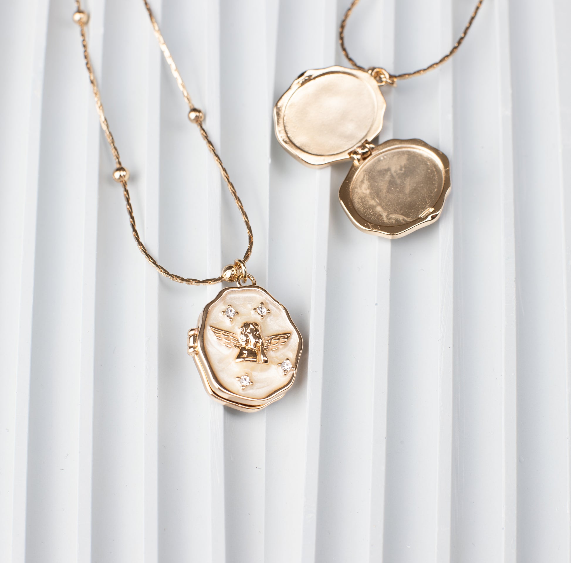 An elegant oval locket necklace with an angel design and sparkling gemstone embellishments, crafted from 18K gold-plated copper. The locket is shown open, revealing a hidden compartment ideal for storing a cherished photo, placed against a modern ridged background to highlight its refined craftsmanship and functionality.
