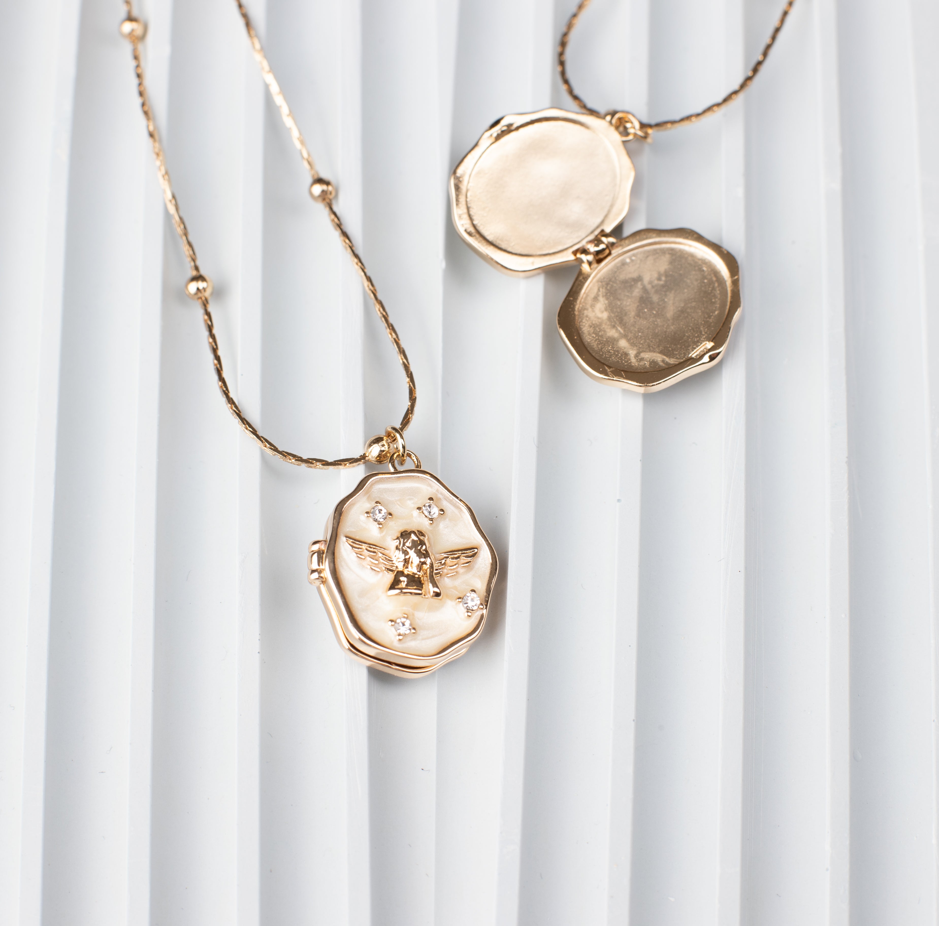 An elegant oval locket necklace with an angel design and sparkling gemstone embellishments, crafted from 18K gold-plated copper. The locket is shown open, revealing a hidden compartment ideal for storing a cherished photo, placed against a modern ridged background to highlight its refined craftsmanship and functionality.