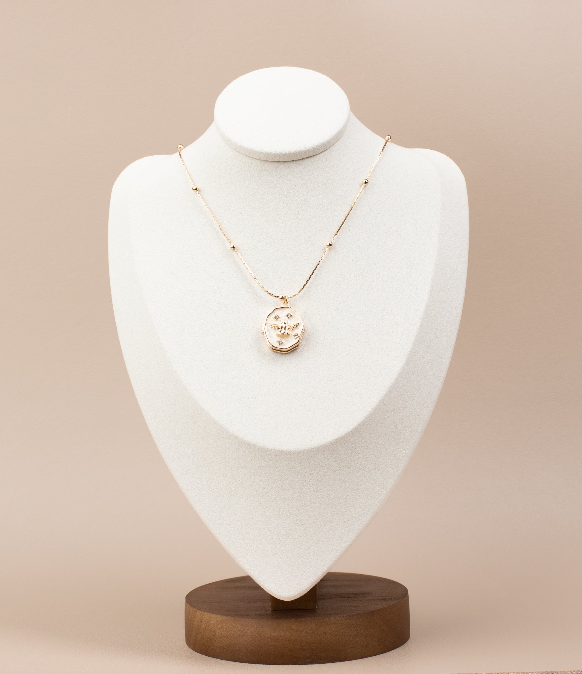 Elegant oval locket necklace with angel design and sparkling gemstone embellishments, crafted from 18K gold-plated copper. The locket can be opened from the side to reveal a compartment inside, perfect for storing a cherished photo. Displayed gracefully on a white velvet bust stand with a sturdy wooden base, highlighting its timeless design and functionality.