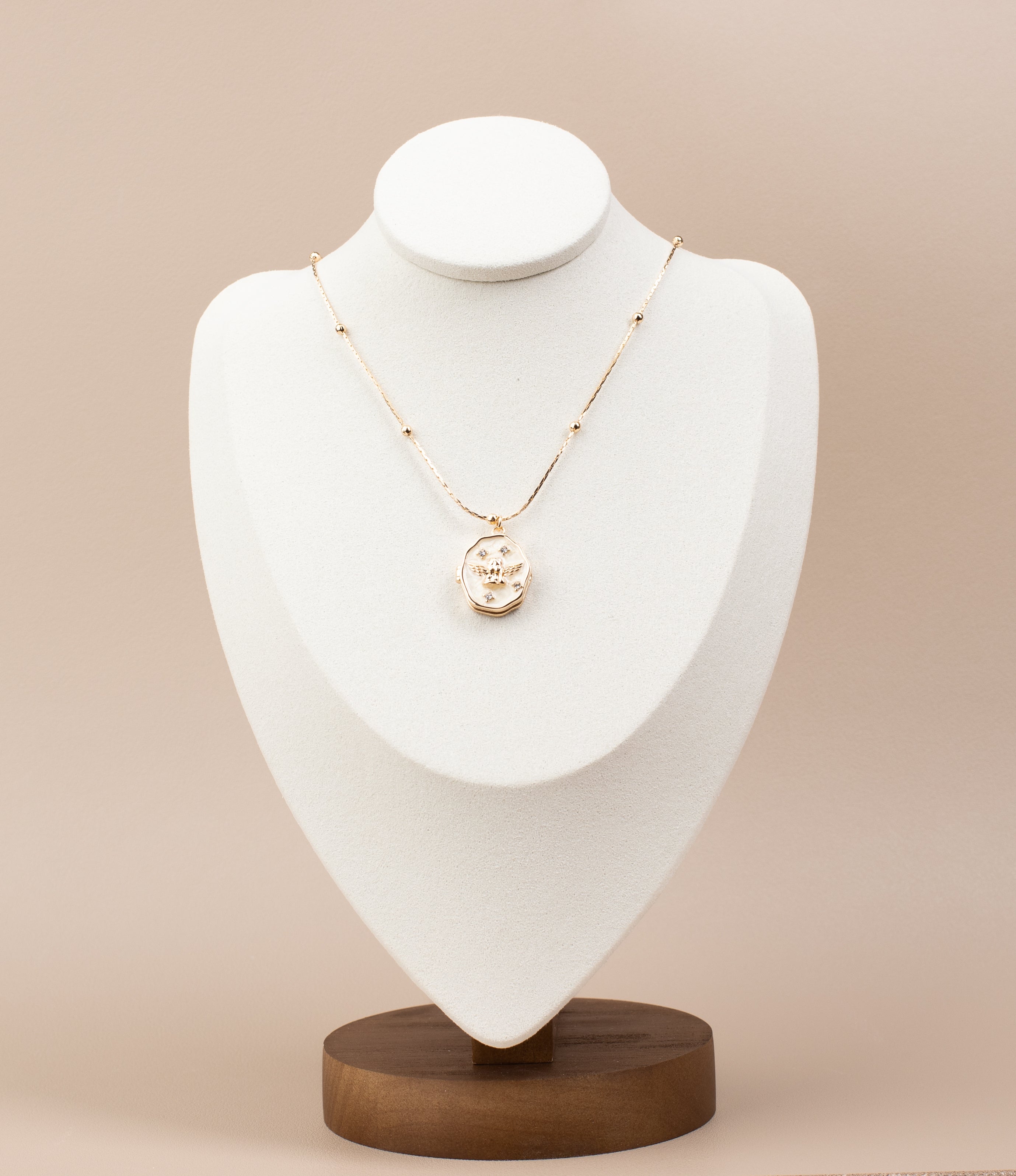 Elegant oval locket necklace with angel design and sparkling gemstone embellishments, crafted from 18K gold-plated copper. The locket can be opened from the side to reveal a compartment inside, perfect for storing a cherished photo. Displayed gracefully on a white velvet bust stand with a sturdy wooden base, highlighting its timeless design and functionality.